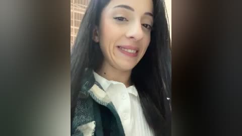 Media: Video of a young woman with long black hair, wearing a white shirt under a green plaid jacket. She has a slight smile, and the background is blurred.