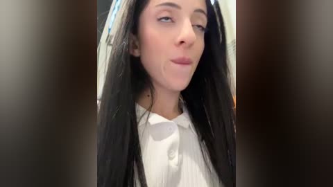 Media: Video of a young woman with long, straight black hair, fair skin, and light makeup, wearing a white blouse. She appears to be indoors with blurred background.