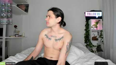 Media: Video of a shirtless man with pale skin and black hair, tattoos on chest and arms, sitting on a bed with white sheets. Room features a \"Follow Us\" neon sign and a potted plant.