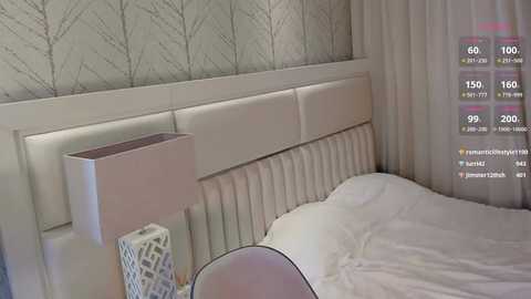 Media: Video of a modern, minimalist bedroom with white furniture, a padded headboard, and a white bedside table with a white lampshade. The wallpaper features a subtle, tree-pattern design. A virtual reality headset is visible on the bed, with digital overlays of social media icons and notifications.