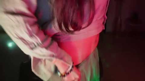 Media: A close-up video of a woman's lower torso and legs, partially obscured by a white, see-through blouse. Her skin tone is light, and she appears to be dancing in a dimly lit room, with a red and green light reflecting on her body.