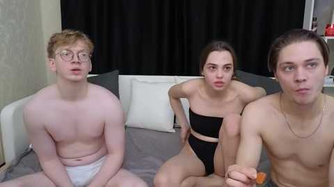 Media: Video of three young people, two women and one man, sitting on a bed. The man, fair-skinned and wearing glasses, sits on the left, shirtless in white briefs. The women, with light skin, wear black lingerie; one with long brown hair and the other with auburn hair.