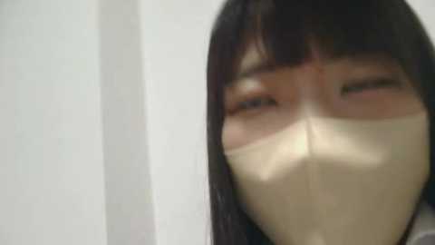 Media: A close-up video of a person wearing a beige face mask, with their eyes barely visible through the mask's slit, against a plain white background.