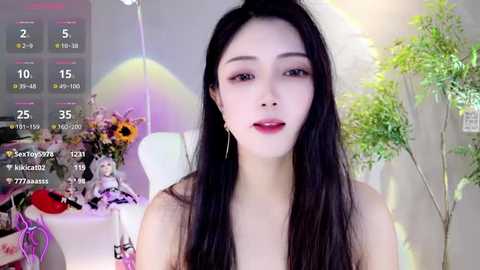 Media: Video of an Asian woman with long black hair, wearing red lipstick, and light makeup, sitting in a room with a white chair, a table with flowers, and a green plant.