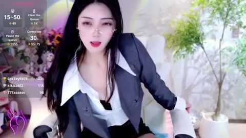 Media: Video of an East Asian woman with long black hair, wearing a black blazer and white shirt, in a room with a plant and colorful background.