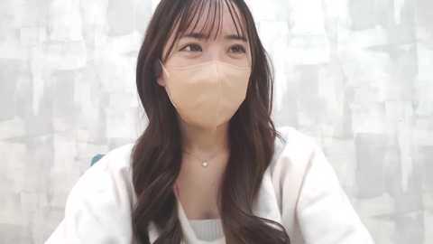 Media: Video of an East Asian woman with long, dark hair, wearing a white mask, white robe, and a delicate necklace, against a blurred, abstract patterned backdrop.