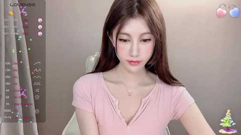 Media: Video of a young Asian woman with long brown hair, wearing a light pink V-neck shirt, sitting in front of a computer screen displaying social media icons and a green chair.