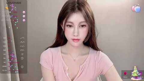 Media: Video of a young East Asian woman with fair skin, long brown hair, and wearing a pink V-neck shirt, in a neutral room with soft lighting, a calendar, and decorative items in the background.