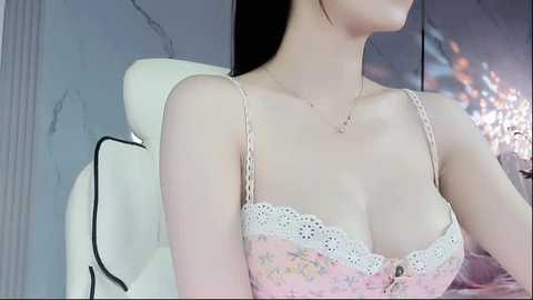 Media: Video of a fair-skinned woman with long black hair, wearing a light pink lace bra, sitting in a modern, white leather chair. The background features a marble wall and a colorful abstract painting.