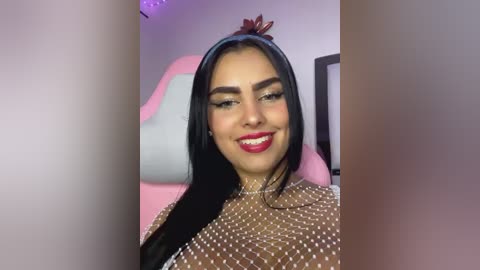 Media: Video of a young woman with long black hair, light skin, and large brown eyes, wearing a sheer top with white polka dots, sitting in a pink gaming chair.