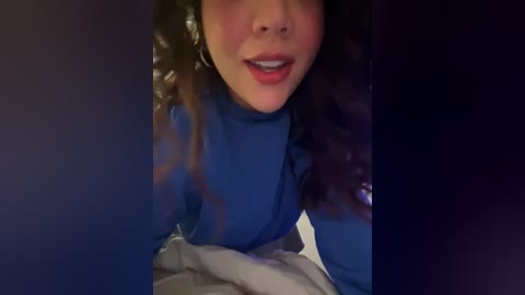 Media: A close-up video of a woman with curly hair, wearing a blue shirt, lying on a bed, partially covered by a blanket, in a dimly lit room.