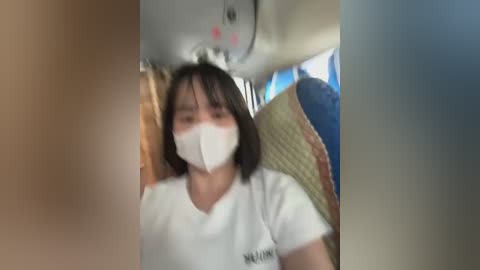 Media: Video of a young woman with short dark hair, wearing a white T-shirt, blue face mask, and sitting on a woven seat inside an airplane. The background shows a blurred window and cabin interior.