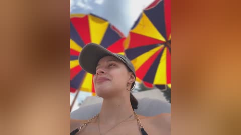 Media: Video of a fair-skinned woman with a tan, wearing a black bikini top and gray cap, looking up with a serene expression. Vibrant, multi-colored umbrellas in the background.