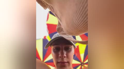 Media: Video of a young man wearing a beige cap and striped shirt, partially obscured by a colorful, geometric umbrella. Background features a bright, multi-colored umbrella.