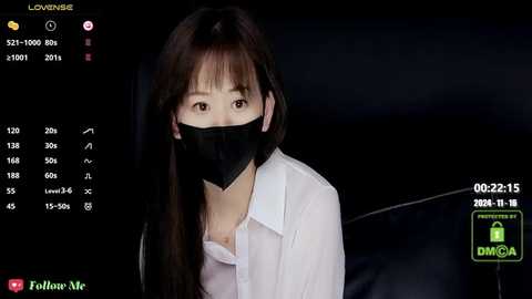 Media: Video of a young East Asian woman with long black hair and fair skin, wearing a black mask and white blouse, set against a dark background.