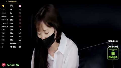 Media: A video of an Asian woman with long black hair, wearing a black face mask, white blouse, and black jacket, seated in a dimly lit room, with a dark background and a digital display showing \"LOVE\" and \"LIVE.\