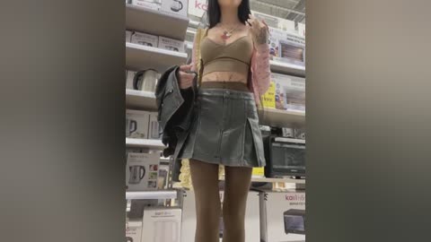 Media: A video of an Asian woman with straight black hair, wearing a beige bra, pink cardigan, and a grey pleated skirt, standing in a disorganized retail store.