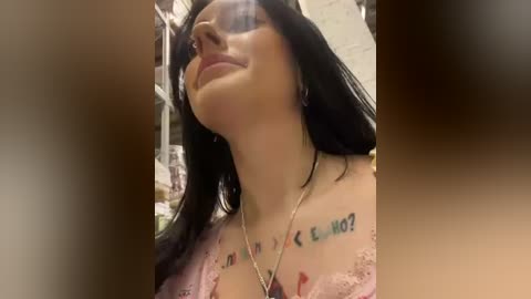 Media: Video of a woman with long black hair, wearing sunglasses and a pink lace top, with tattoos on her shoulders, smiling slightly.