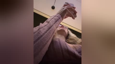 Media: A video of a woman taking a selfie from a low angle, wearing a pink sweater, standing in a room with beige walls and a black speaker.