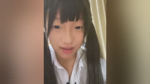 Media: A close-up video of a young East Asian girl with long black hair, wearing a white collared shirt. She has a calm expression, and the background is a plain beige wall.