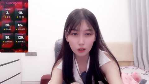 Media: A video of a young East Asian woman with long black hair, wearing a white top, sitting on a bed with a pink pillow, in a minimalist bedroom with a digital display showing \"LOVEYOUNG\" and a chart.