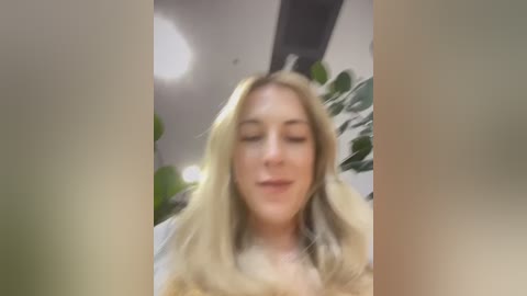 Media: Video of a smiling blonde woman with long hair, wearing a white top, in a modern indoor setting with green plants. The image is slightly blurry.