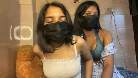 Media: Video of two women with medium brown skin, wearing black face masks, sitting on a couch with beige walls. One wears a white lace top, the other a blue off-shoulder top.