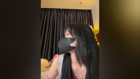 Media: Video of a young woman with long black hair, wearing a black face mask and a light blue top, sitting in a dimly-lit room with black curtains and plush toys.