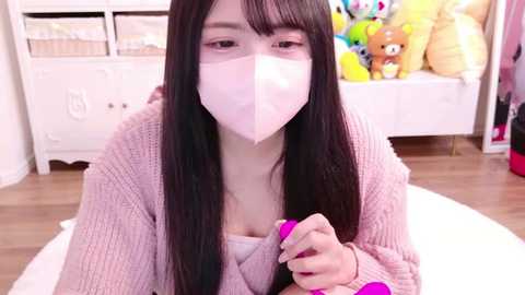 Media: Video of an Asian woman with long black hair, wearing a pink face mask and a knitted sweater, holding a pink hairbrush in a cozy, pastel-colored bedroom with stuffed animals.