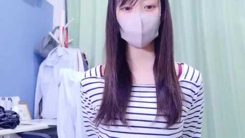 Media: Video of an Asian woman with long, straight black hair, wearing a gray surgical mask and a white and black striped long-sleeve shirt, standing in a bright, clean bathroom with light green curtains and white walls.