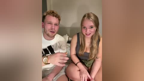 Media: Video of a young couple, a blonde woman in a grey tank top and khaki shorts, and a curly-haired man in a white shirt, sitting on a bed.