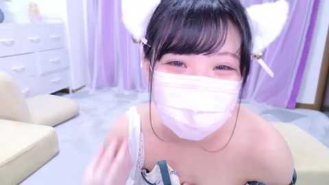 Media: A young Asian woman with fair skin, wearing a white face mask, white cat ears, and a black lace bra, sits on a beige couch in a softly lit, pastel-colored room.