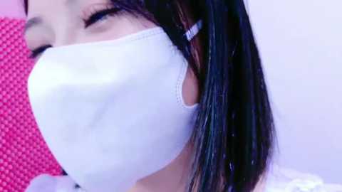 Media: A close-up video of an Asian woman with straight black hair, wearing a white surgical mask, partially visible face, and a pink textured background.