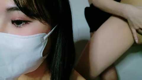 Media: Video of a young Asian woman with straight, dark hair wearing a white surgical mask. She's close to another person, possibly a man, who is wearing black shorts. The background is blurred, focusing on their faces and upper bodies.