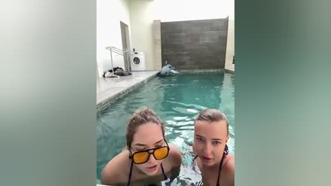 Media: Video of two young women in a swimming pool, one with sunglasses, both smiling. Pool surrounded by light-colored walls and a dark wooden door, clothes and a drying rack in the background.