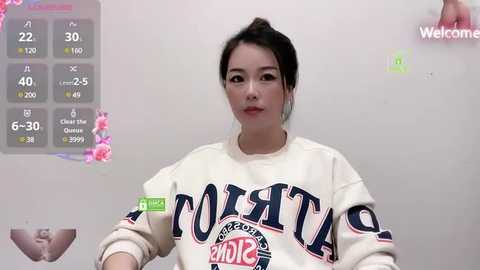 Media: Video of a young Asian woman with fair skin, dark hair tied back, wearing a white sweatshirt with \"PANTHERS\" and \"WELCOME TO THE GAME\" printed in blue and red. Background includes a digital health tracker displaying vital signs.