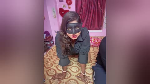 Media: Video of a dark-haired woman in a black mask, kneeling on a patterned bedspread, in a pink room with heart decorations and red curtains.