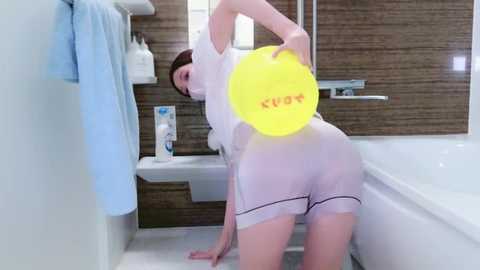 Media: Video of a woman with short brown hair, wearing a sheer white dress and panties, bending over in a modern bathroom with tiled walls, a white sink, and a bathtub.