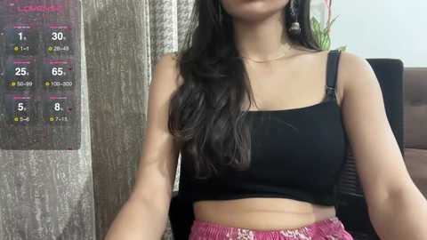 Media: Video of a young woman with long black hair, wearing a black tank top and pink high-waisted shorts, sitting in front of a calendar and a plant.