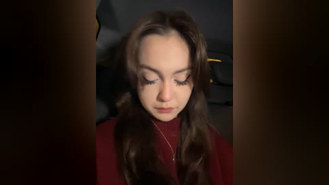 Media: Video of a young woman with long, wavy brown hair, wearing a red sweater, sitting in a dimly lit car. Her eyes are closed, and she appears serene, with a soft focus background.