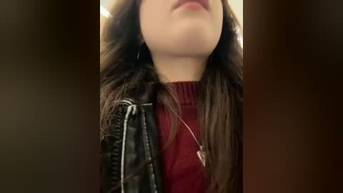 Media: Video of a young woman with long brown hair, wearing a black leather jacket over a red turtleneck, looking downward, indoors with blurred background.