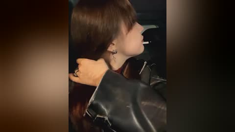 Media: Video of a young woman with long brown hair, wearing a black leather jacket, leaning against a car with a lit cigarette in her mouth, set in a dimly lit, urban background.