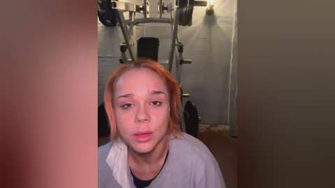 Media: A video of a young woman with light skin and shoulder-length, reddish-brown hair, wearing a grey T-shirt, sitting on a weight bench in a dimly lit gym with white walls and equipment in the background.