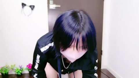 Media: Video of a young Asian woman with shoulder-length black hair, wearing a black jacket, leaning over a desk, indoors, with a white wall and a pink plant in the background.