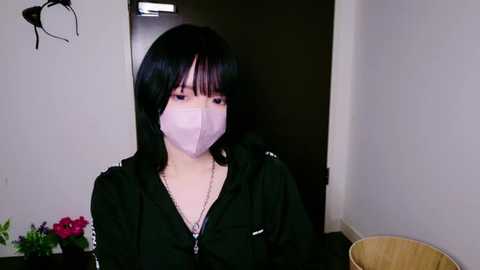 Media: Video of an East Asian woman with straight black hair, wearing a black hoodie, pink face mask, and a silver necklace, standing in a dimly lit room with a wooden chair and red flowers in the background.