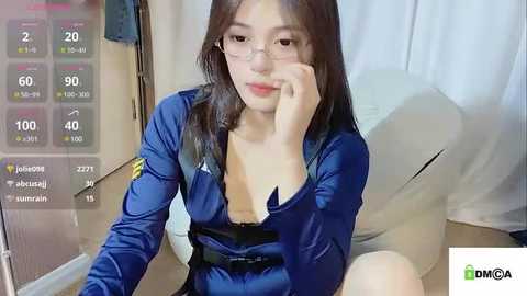 Media: A video of a young Asian woman with long black hair, wearing a blue police uniform with a low neckline, sitting on a bed, adjusting her glasses, in a bedroom.