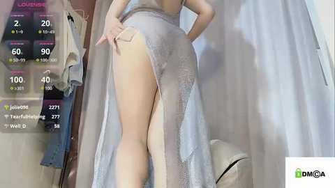 Media: Video of a fair-skinned woman wearing a sheer, silver lace dress with a high slit, revealing her bare buttocks and thighs. She stands in a dimly lit room with a closet and a white curtain in the background.