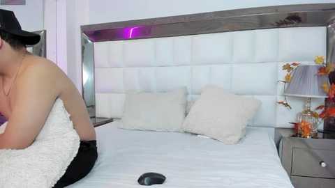 Media: A video of a topless woman with fair skin, wearing black lingerie, kneeling on a white bed with a white headboard. The background features a modern bedroom with white walls and a purple accent light.