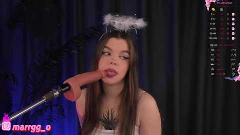 Media: Video of a young woman with long brown hair, wearing a white feathered halo, purple lipstick, and a feathered necklace, blowing into a microphone. Background shows black curtains and a live stream interface.