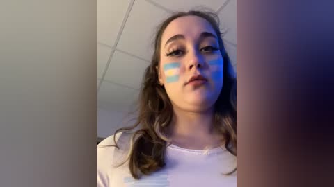 Media: Video of a young woman with light skin, brown hair, wearing a white shirt, with blue and pink paint on her face resembling the Pride flag, taken indoors.